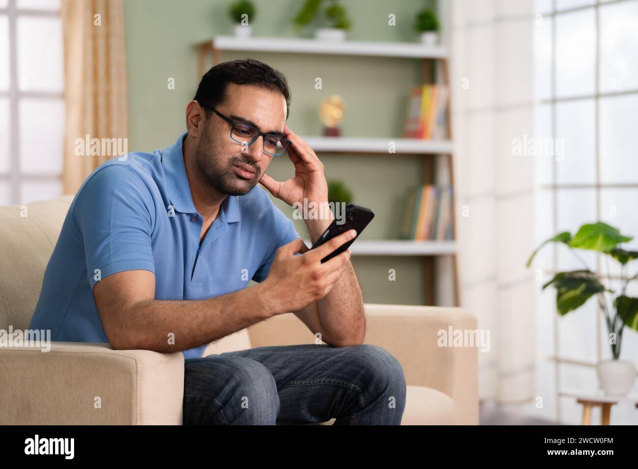 Schockierter indischer Mann bekam schlechte Nachrichten, als er zu Hause das Handy auf dem Sofa benutzte - Konzept von finanzieller Not, Kreditstornierung und unsicherer Zukunft Stockfoto