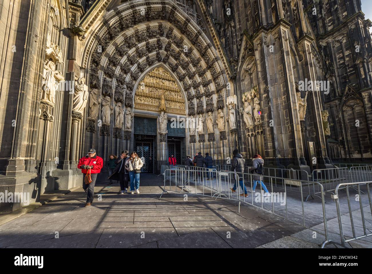 Besucher stehen am reich verzierten Hauptportal des Kölner Doms in Köln, Nordrhein-Westfalen, Deutschland, mit Barrieren, die die Eingangsschlange lenken. Steinfiguren von Heiligen säumen das Portal unter einem spitzen Tympanon. Der Bau der gotischen Kathedrale begann 1248 und wurde nach einer jahrhundertelangen Unterbrechung erst 1880 fertiggestellt. Mit einer Höhe von 157 Metern sind die beiden Türme die höchsten Zwillings­türme einer Kirchenfassade weltweit; die Kathedrale ist seit 1996 UNESCO-Weltkulturerbe. Stockfoto