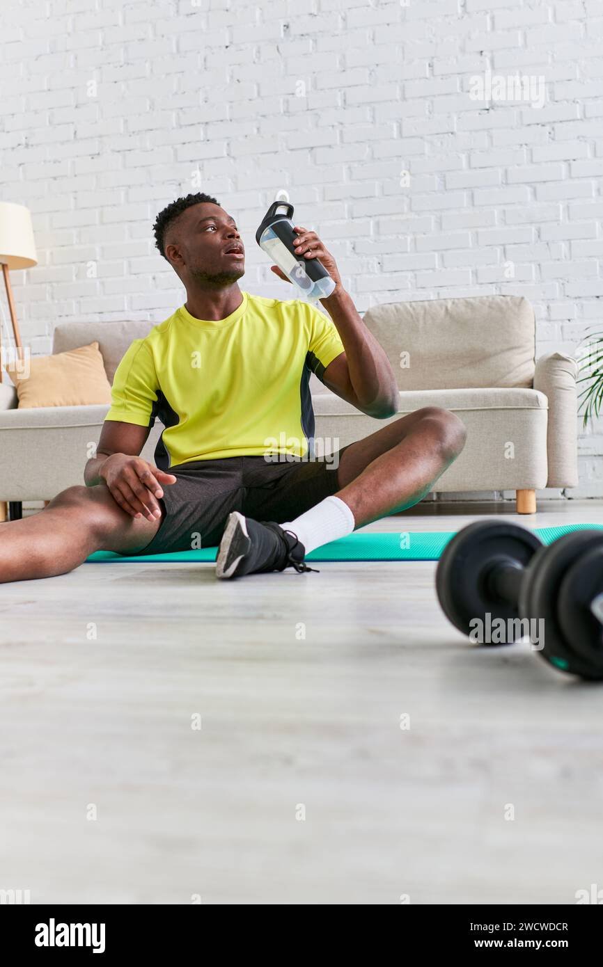 Durstiger afroamerikanischer Mann in Sportbekleidung, der auf einer Fitnessmatte mit einer Sportflasche Süßwasser sitzt Stockfoto