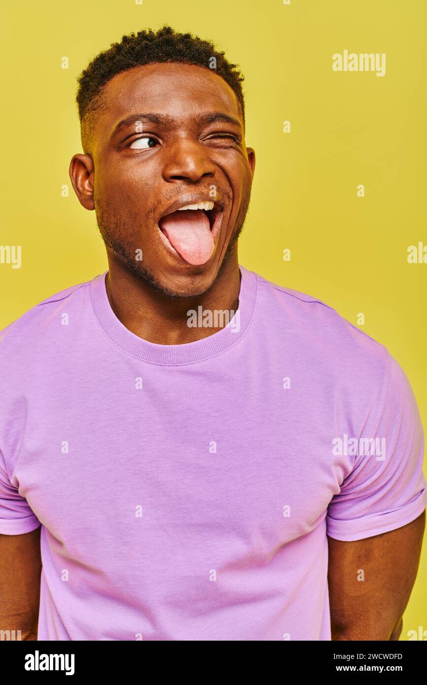 Lustiger afroamerikanischer Mann in lila T-Shirt, der die Zunge herausragt und vor gelbem Hintergrund wegschaut Stockfoto