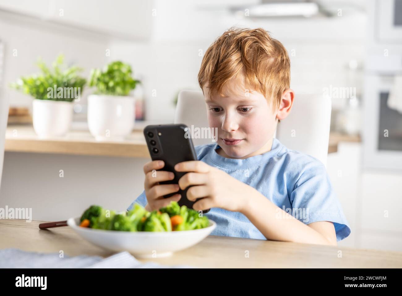 Blick auf den schicken, süßen Jungen, der vor dem Mittagessen in der Küche zu Hause ein Smartphone verwendet. Stockfoto