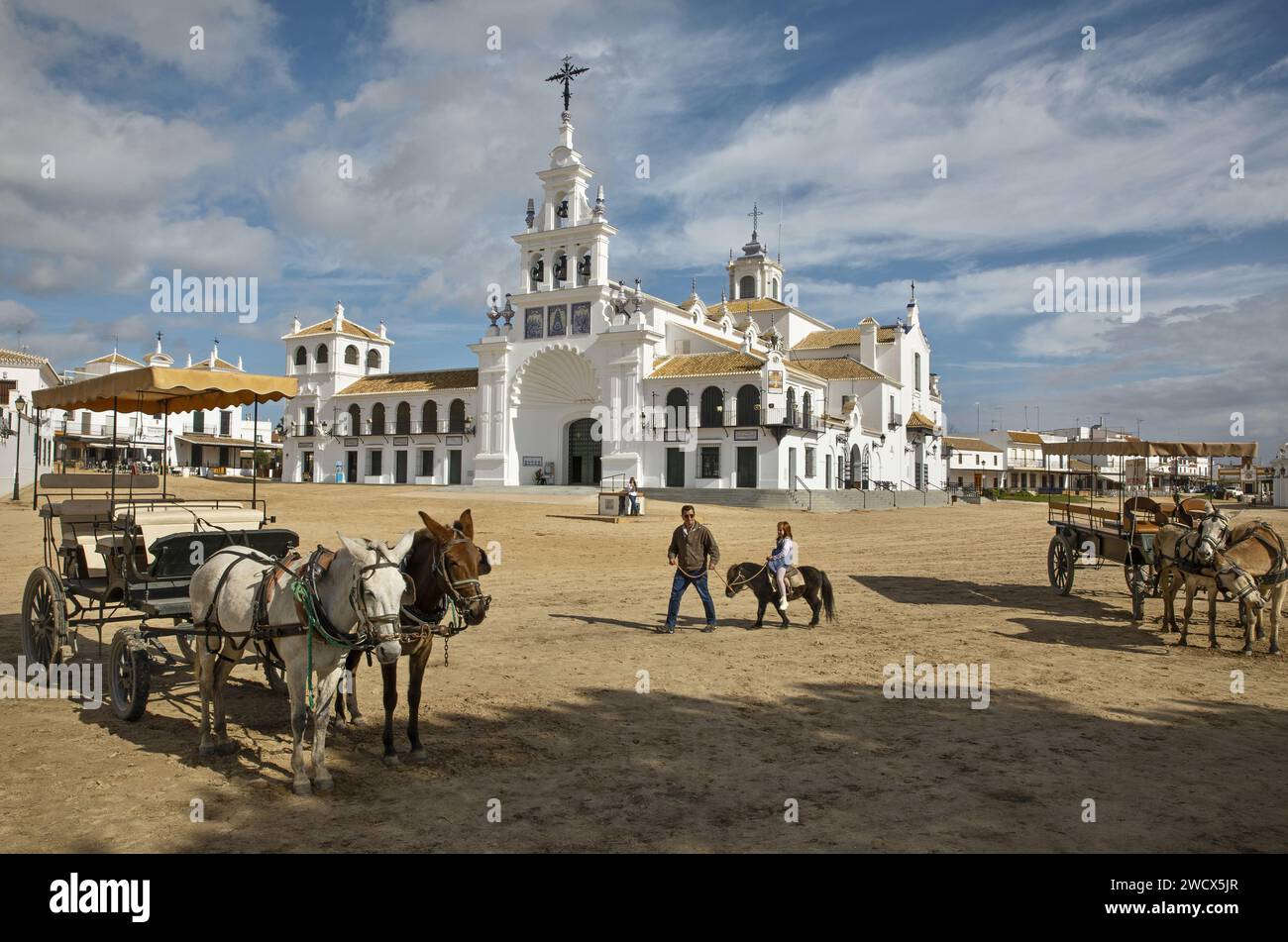 Spanien, Andalusien, El Rocío, Mann, der seine kleine Tochter auf einem Pony in einer Sandstraße mit Pferdewagen vor der Eremitage Nuestra señora del Rocío führt Stockfoto
