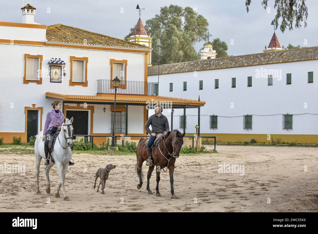 Spanien, Andalusien, El Rocío, junges Paar zu Pferd mit Hund an der Seite in einer Sandstraße gesäumt von Hermandaden, den Häusern katholischer Bruderschaften Stockfoto
