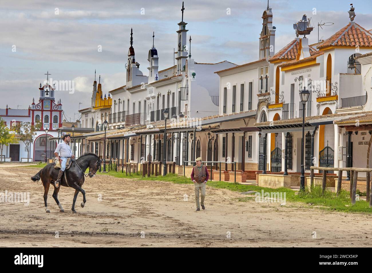 Spanien, Andalusien, El Rocío, Reiter auf einem schwarzen Pferd neben einem alten Mann mit Baskenmütze in einer Sandstraße, die von Hermandaden gesäumt ist, den Häusern katholischer Bruderschaften Stockfoto