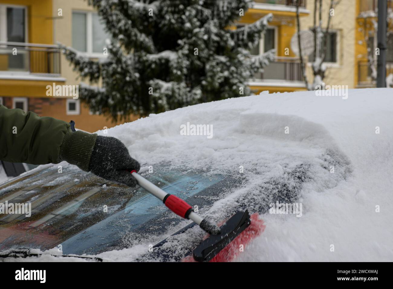 Ein Typ in Jacke, Hut und Handschuhen fegt das Auto nach einem Schneesturm mit einem kleinen roten Besen. Stockfoto
