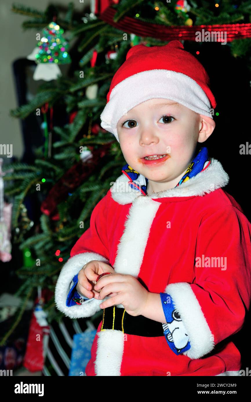 Der Junge, der in einem Weihnachtsmann-Anzug verkleidet ist, macht eine Pause von der Eröffnung der Geschenke und isst am Weihnachtsmorgen Schokolade. Stockfoto