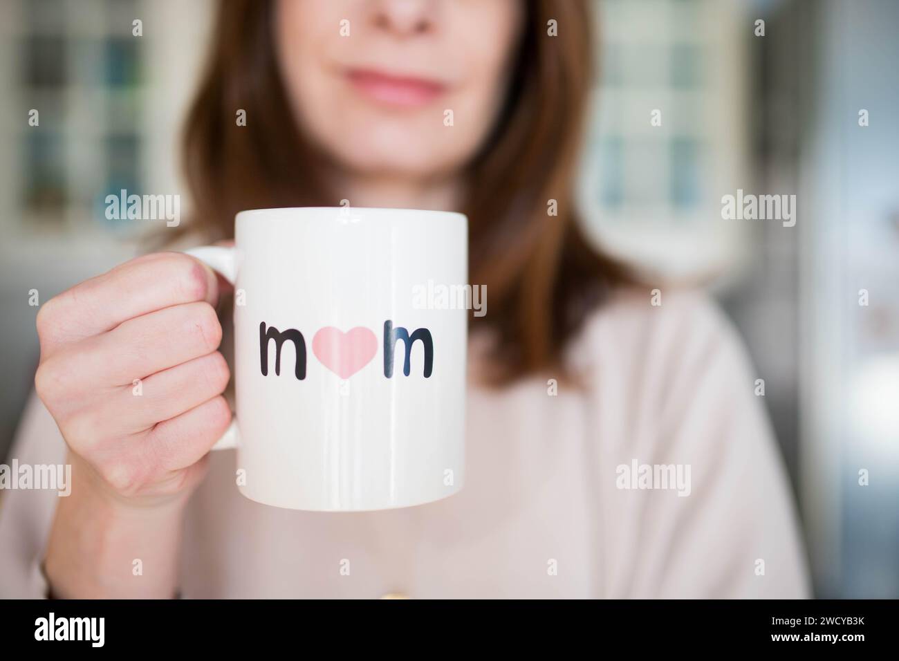 Eine weiße Frau, die glücklich darüber lächelt, Mutter zu sein, hält einen Muttertagsbecher mit 'Mutter' drauf. Rosafarbener, luftiger Hintergrund. Stockfoto