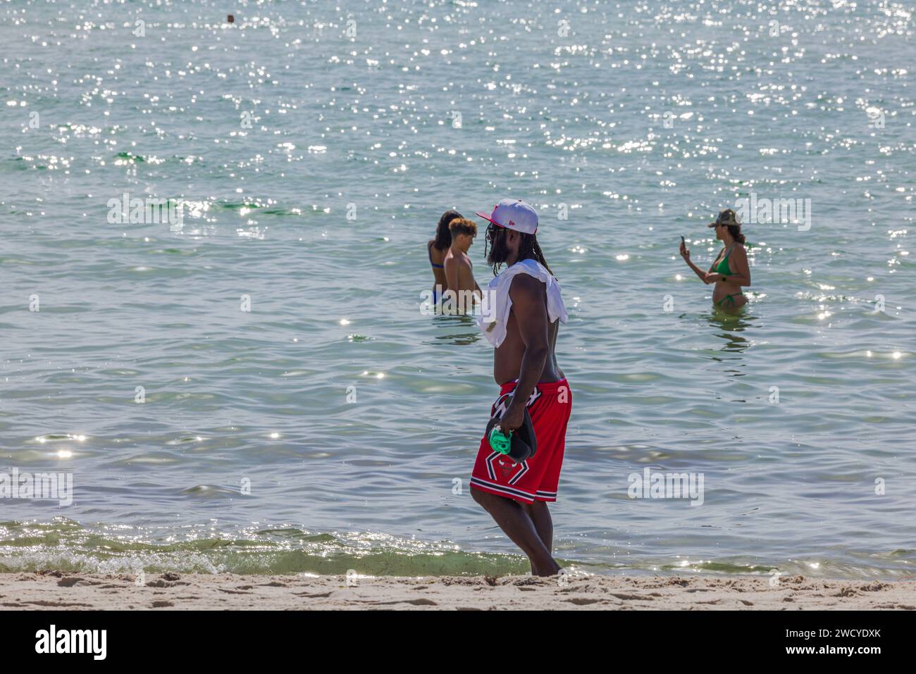 Ein hübscher junger Afroamerikaner mit sportlichem Aufbau, in roten Badeshorts und einer weißen Mütze, schlendert entlang der Küste von Miami Beach auf einer Stockfoto