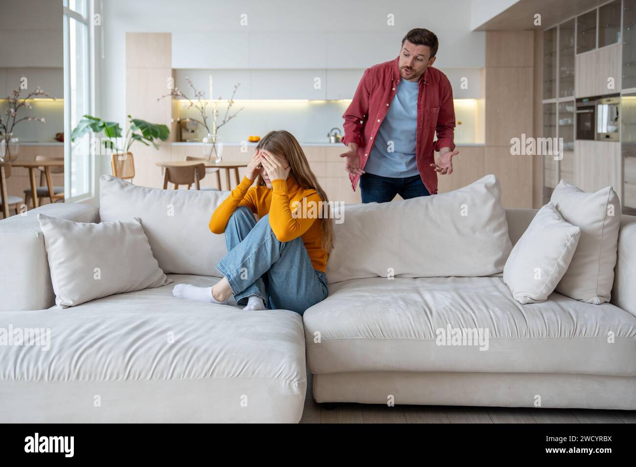 Ein wütender Mann schreit die Frau an, die auf dem Sofa sitzt und das Gesicht mit Händen bedeckt, Konflikt, emotionale Not Stockfoto