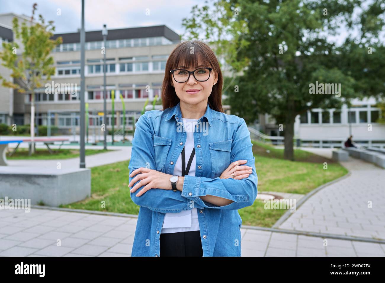 Selbstbewusste Lehrerin mittleren Alters mit gekreuzten Armen in der Nähe des Schulgebäudes Stockfoto