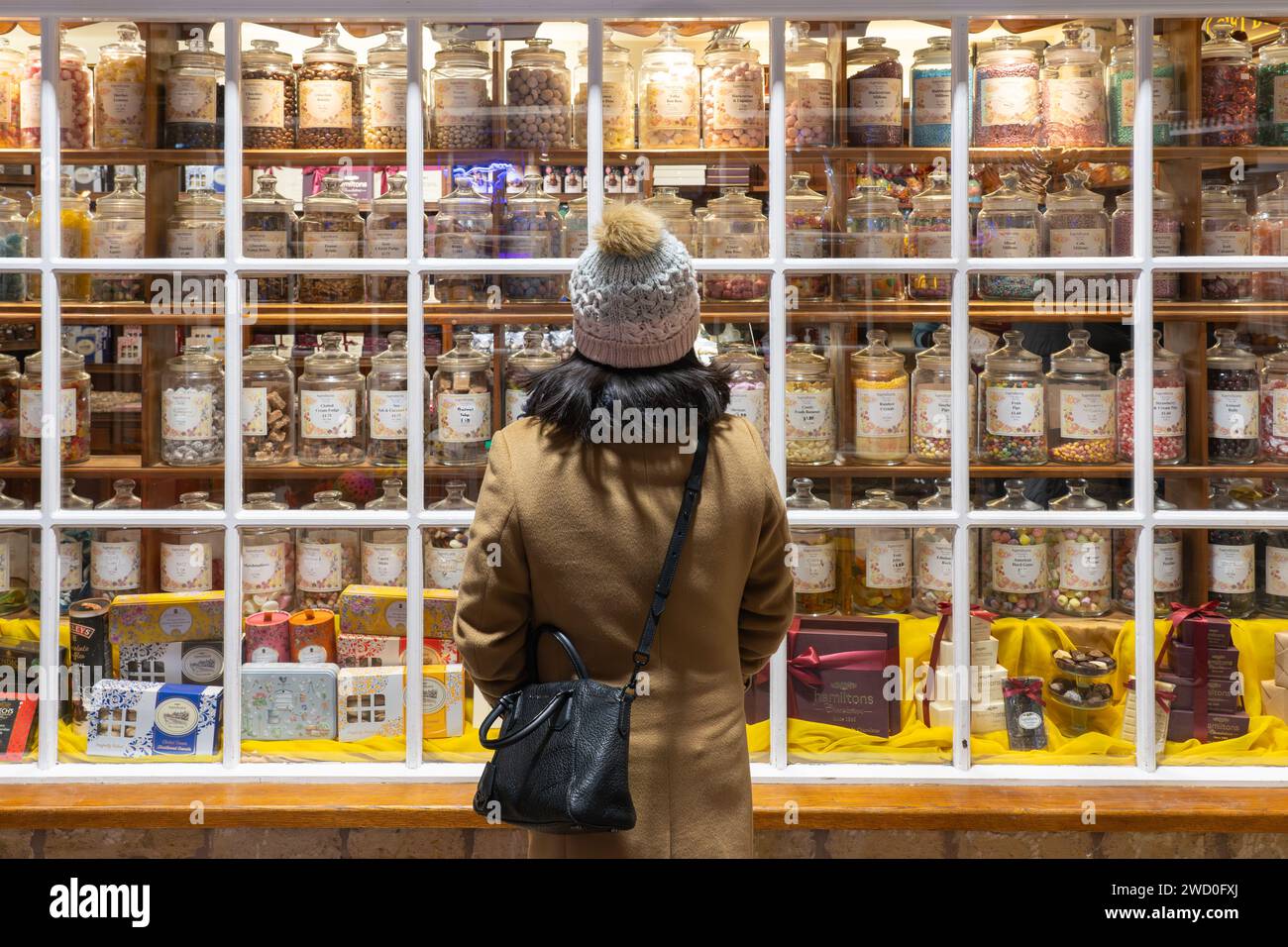 Eine Frau, die im Winter einen Wackelhut trägt, schaut in das Fenster von Hamiltons of Broadway, einem süßen Laden mit traditionellen englischen Süßwaren. England Stockfoto