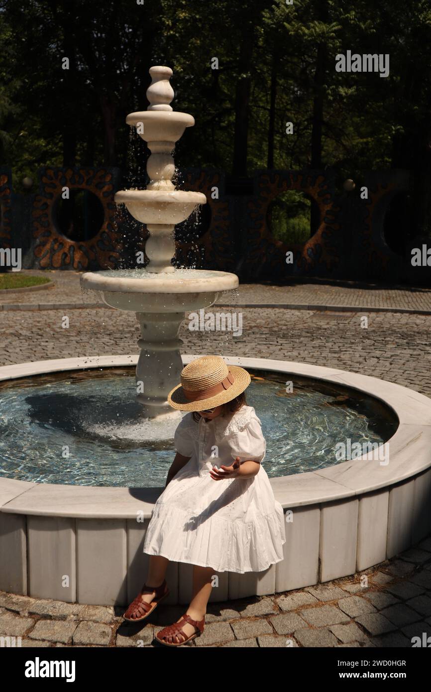 Eine vertikale Aufnahme eines entzückenden Mädchens in einem Kleid, das einen ruhigen Moment an einem wunderschönen Brunnen genießt Stockfoto