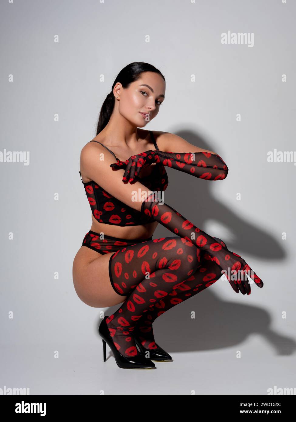 Junge sexy Brünette Frau in High Heels, die in roten und schwarzen Dessous sitzt, lange Strümpfe und Handschuhe mit Lippenmuster vor grauer Wand und er Stockfoto