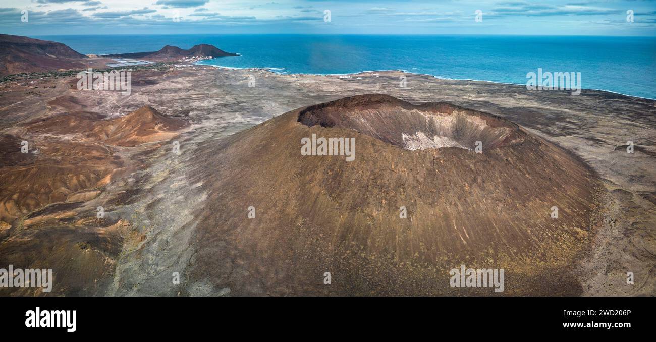 Luftaufnahme einer vulkanischen Küstenlandschaft (Vulkan Viana, Sao Vicente) mit einem markanten Krater im Vordergrund, karger Vegetation und einer kleinen Ville Stockfoto