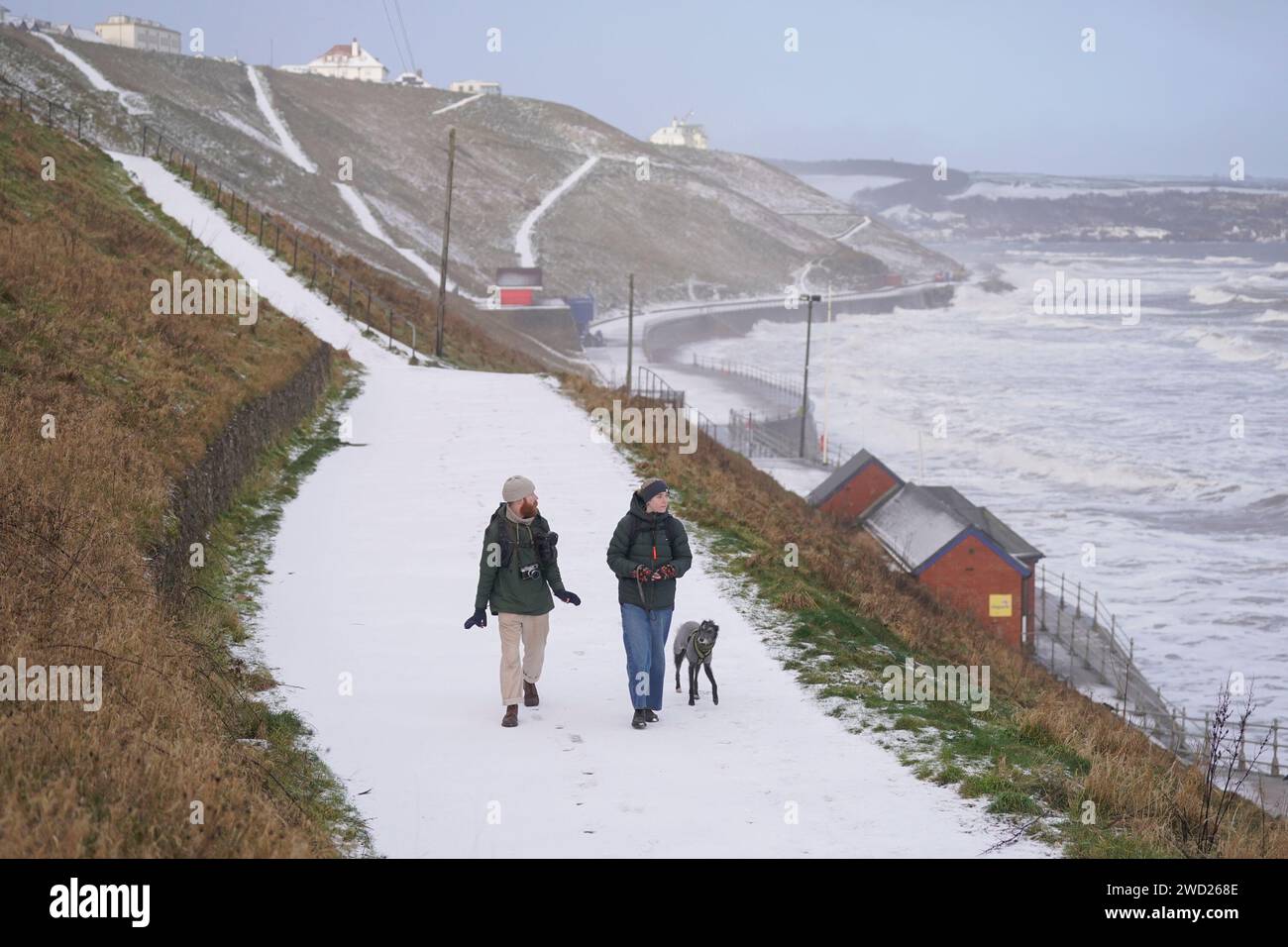 Menschen gehen mit einem Hund auf einem schneebedeckten Weg in Whitby. Hunderte von Schulen sind geschlossen, einige für einen vierten Tag, da in vielen Teilen des Landes Wetterwarnungen einschließlich eines gelben Alarms bestehen bleiben. Bilddatum: Donnerstag, 18. Januar 2024. Stockfoto