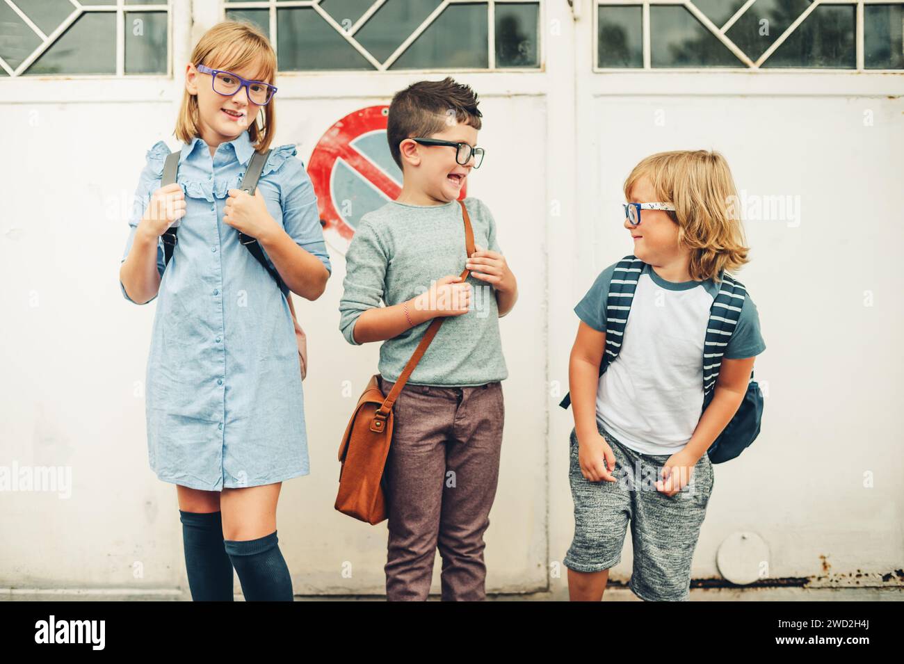 Eine Gruppe von drei lustigen Kindern, die Rucksäcke tragen und zurück zur Schule gehen. Mädchen und Jungs mit Brille, die draußen posieren Stockfoto