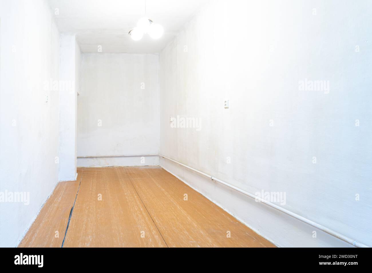 Leeres Zimmer mit Gebrauchsspuren, weiße Wände, die neu gestrichen werden müssen, ein alter Holzboden mit sichtbaren Schäden und eine einfache Deckenleuchte Stockfoto