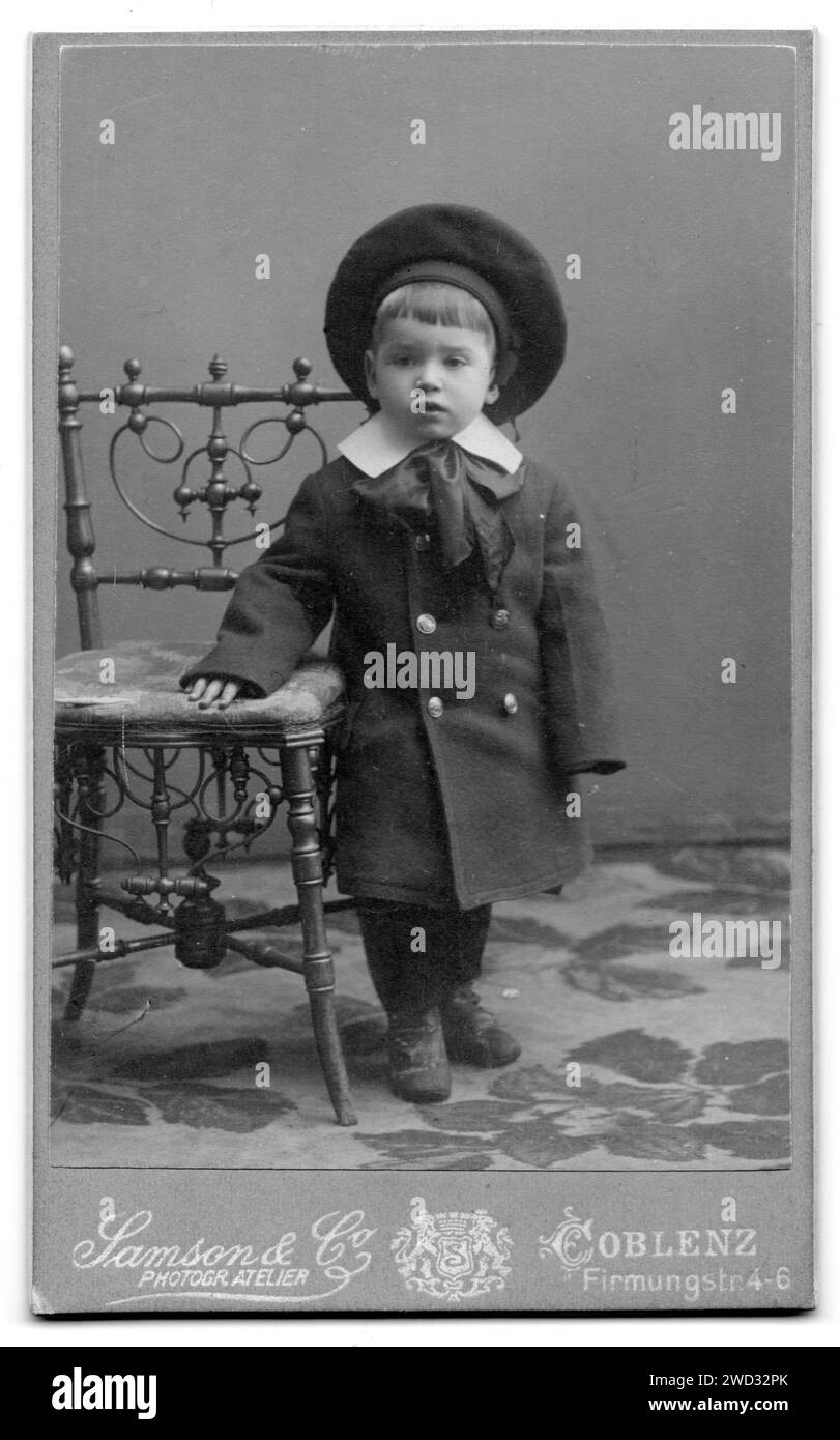 Studio-Porträt eines kleinen Jungen in einem Mantel, mit einer Schleife um den Hals und einer Baskenmütze, der neben einem Stuhl steht. Der Junge heißt Hans Bach6, auf dem er geboren wurde Stockfoto