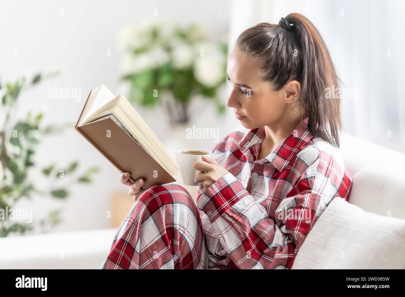 Die schöne Brünette im Pyjama liest ein Buch, das auf dem Sofa im Wohnzimmer sitzt. Stockfoto
