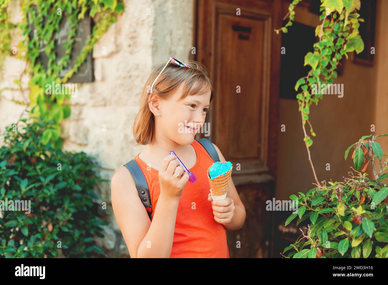 Niedliches kleines Mädchen, das draußen blaues Eis isst, oranges Hemd und Rucksack trägt und mit Kindern reist Stockfoto