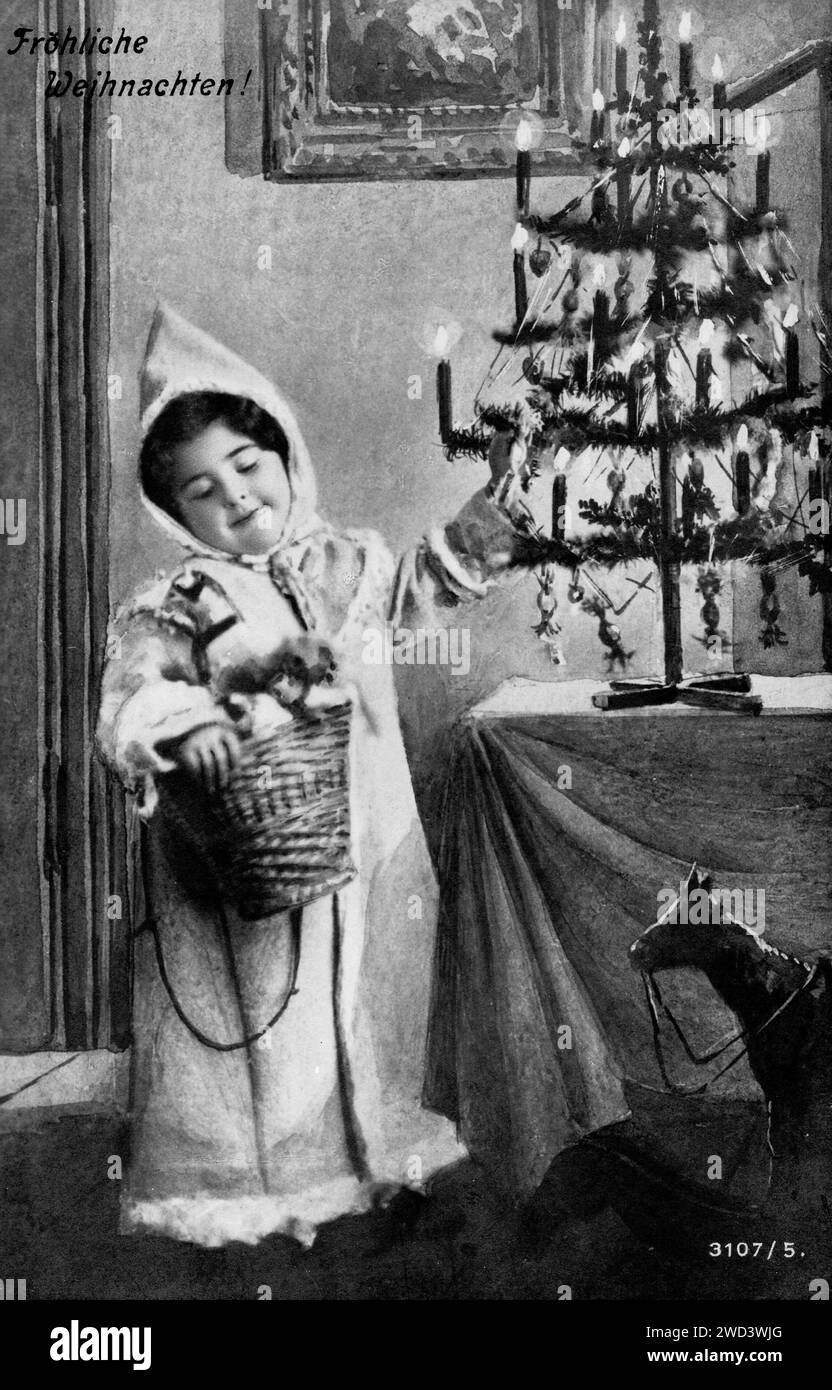 Eine antike Postkarte zeigt ein Mädchen mit einem mit Geschenken gefüllten Korb neben einem Tisch, auf dem ein Weihnachtsbaum steht. Inschrift auf Deutsch: Mer Stockfoto