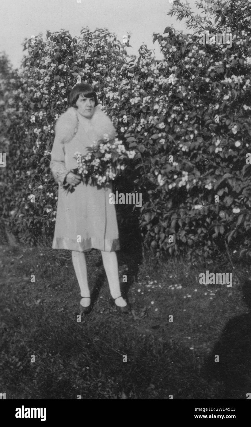 Ein antikes Foto zeigt eine Frau in einem weißen Mantel mit einem Pelzkragen, die in einem blühenden Garten mit einem Blumenstrauß in den Händen steht. Deutschland. Datum unbekannt, ci Stockfoto