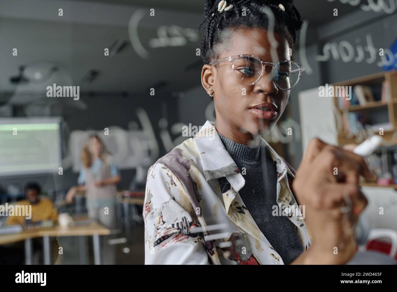 Eine Coderin schreibt nachdenklich im Büro an einer Glaswand, ihre Kollegen arbeiten am Hintergrund Stockfoto