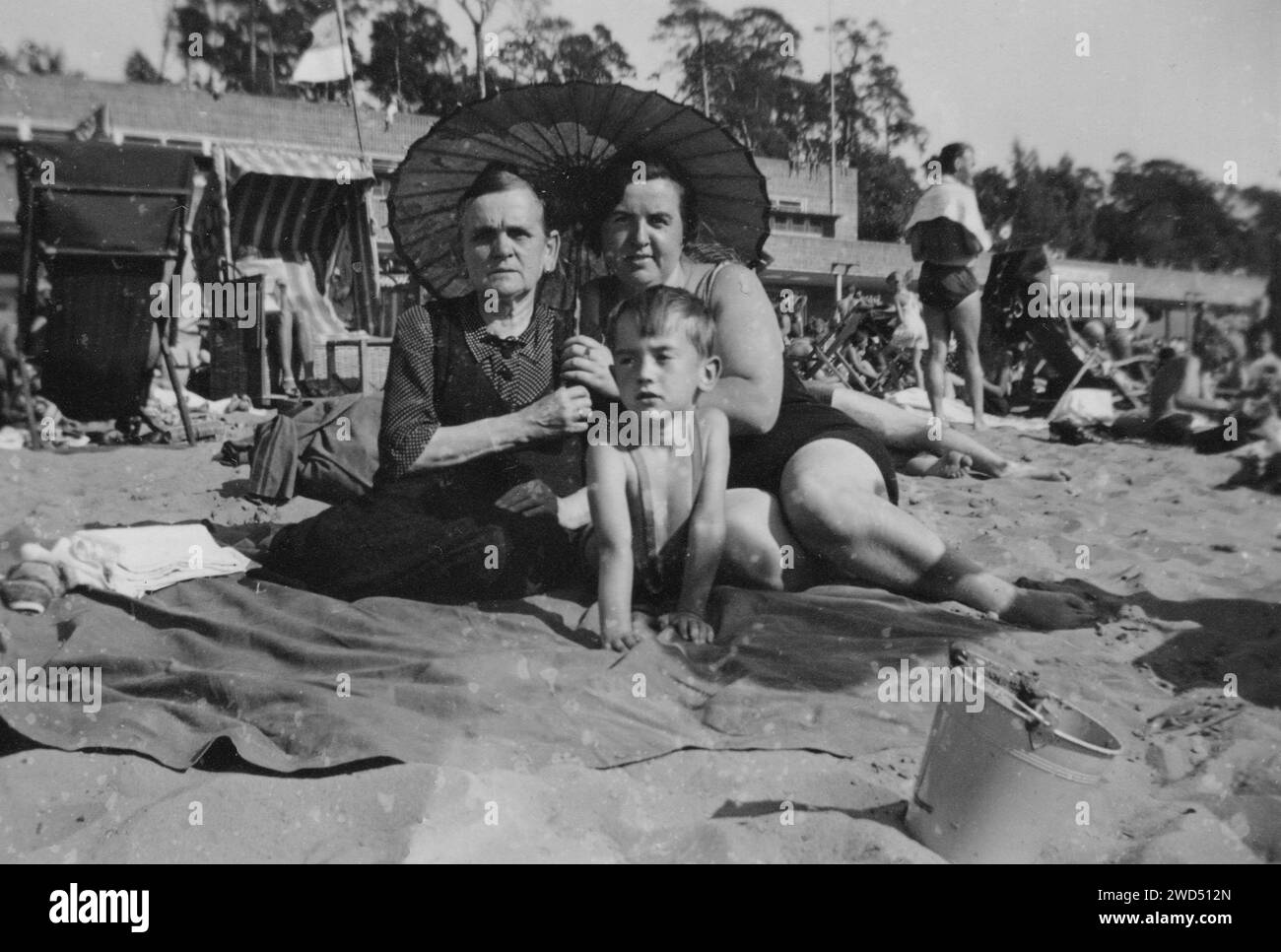 Ein altes Foto zeigt zwei kaukasische Frauen, eine ältere in einem Kleid und die andere in einem Badeanzug, und einen kleinen Jungen, der unter einem Sonnenschirm am Strand posiert Stockfoto