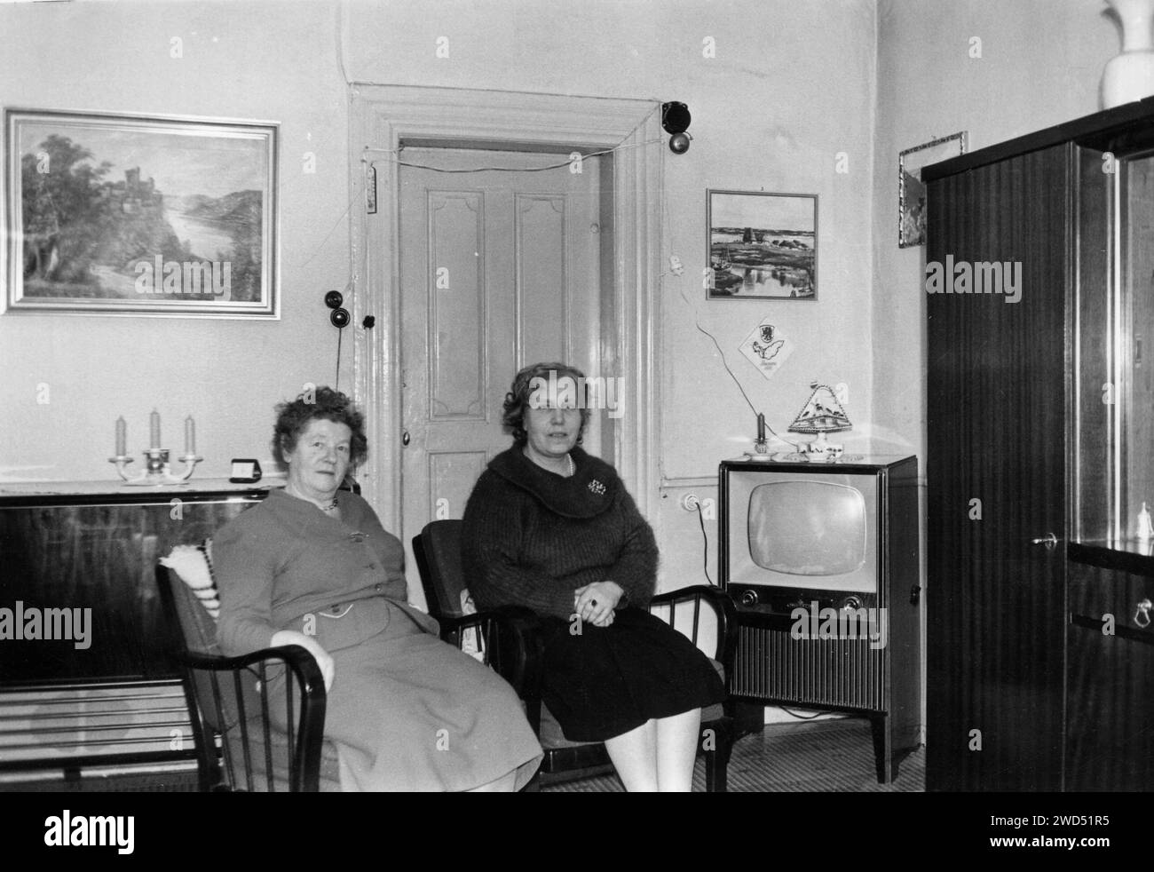 Ein antikes Foto zeigt zwei ältere kaukasische Frauen, die auf den Stühlen im Inneren des Wohnzimmers sitzen, in dem sich ein Fernseher befindet Stockfoto