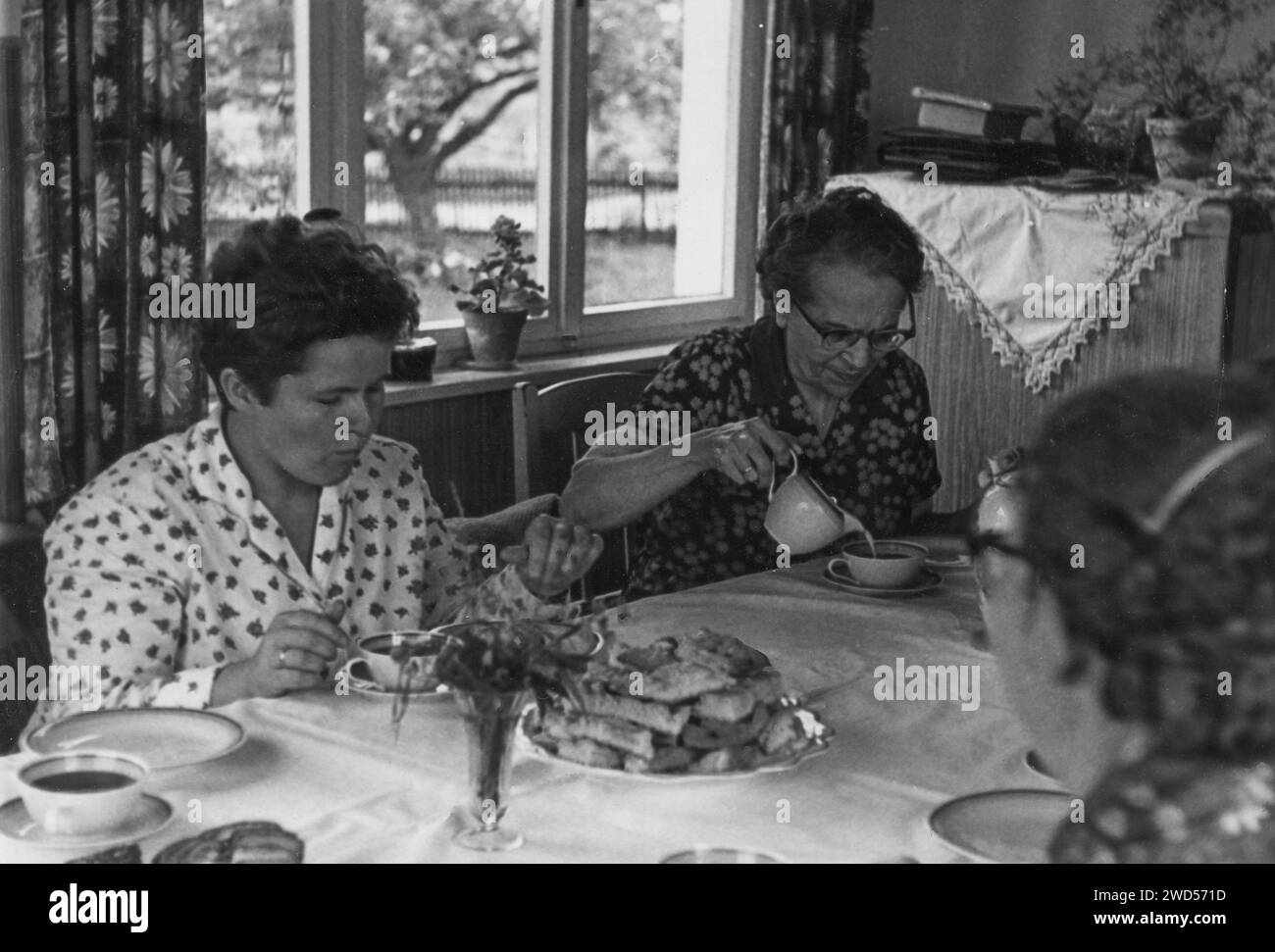 Ein antikes Foto zeigt drei Frauen, die Tee mit hausgemachten Keksen an einem Tisch im Wohnzimmer trinken. Deutschland. Datum unbekannt, um die 1950er Jahre Stockfoto