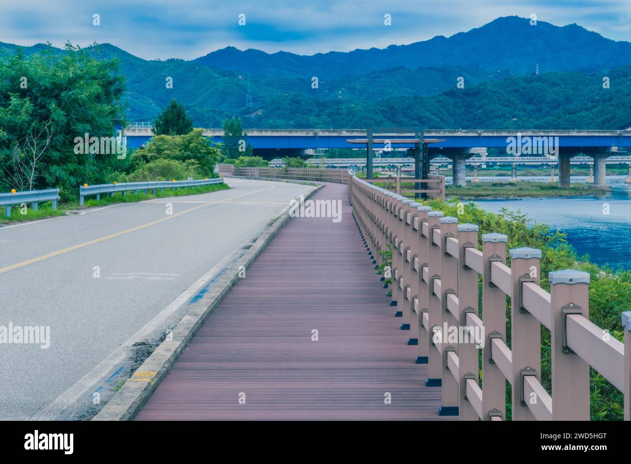 Hölzerner Bürgersteig entlang einer Landstraße neben dem Fluss mit Autobahnbrücke und Bergen unter bewölktem Himmel im Hintergrund, Südkorea Stockfoto