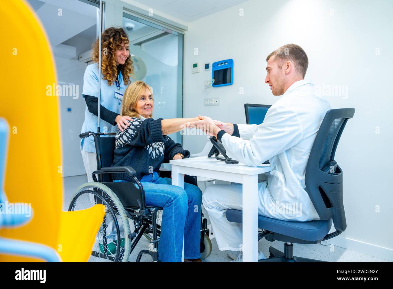 Arzt schüttelt eine reife Frau im Rollstuhl in einem Krankenhauszimmer mit Schreibtisch und Computer Stockfoto