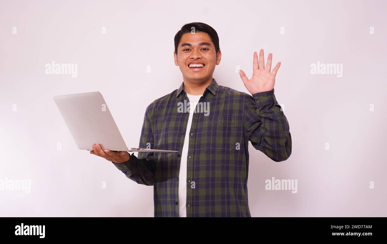 Asiatischer Schüler winkt die Hand, während er den Laptop vor weißem Hintergrund hält Stockfoto
