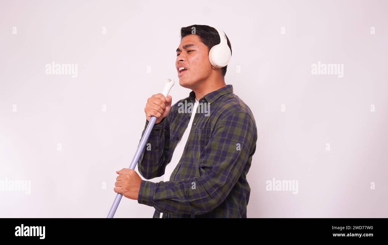 Asiatischer Mann, der Musik mit Kopfhörern hört und auf Besen vor weißem Hintergrund singt Stockfoto
