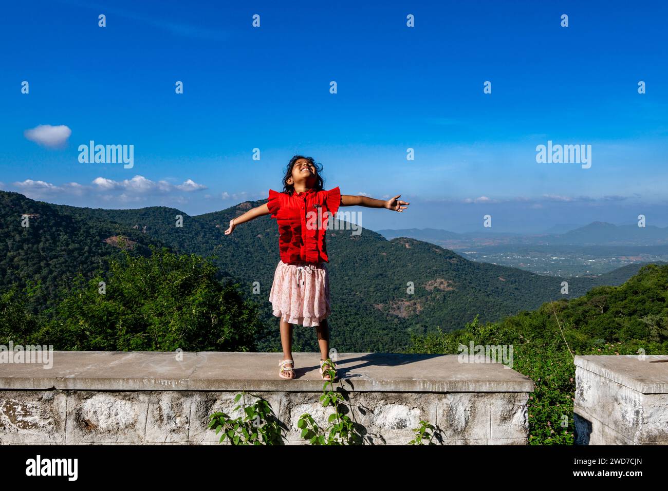 Ein kleines Kind steht triumphierend auf einem Berggipfel, die Arme zum Himmel gestreckt und die Welt als ihre eigene bezeichnet Stockfoto