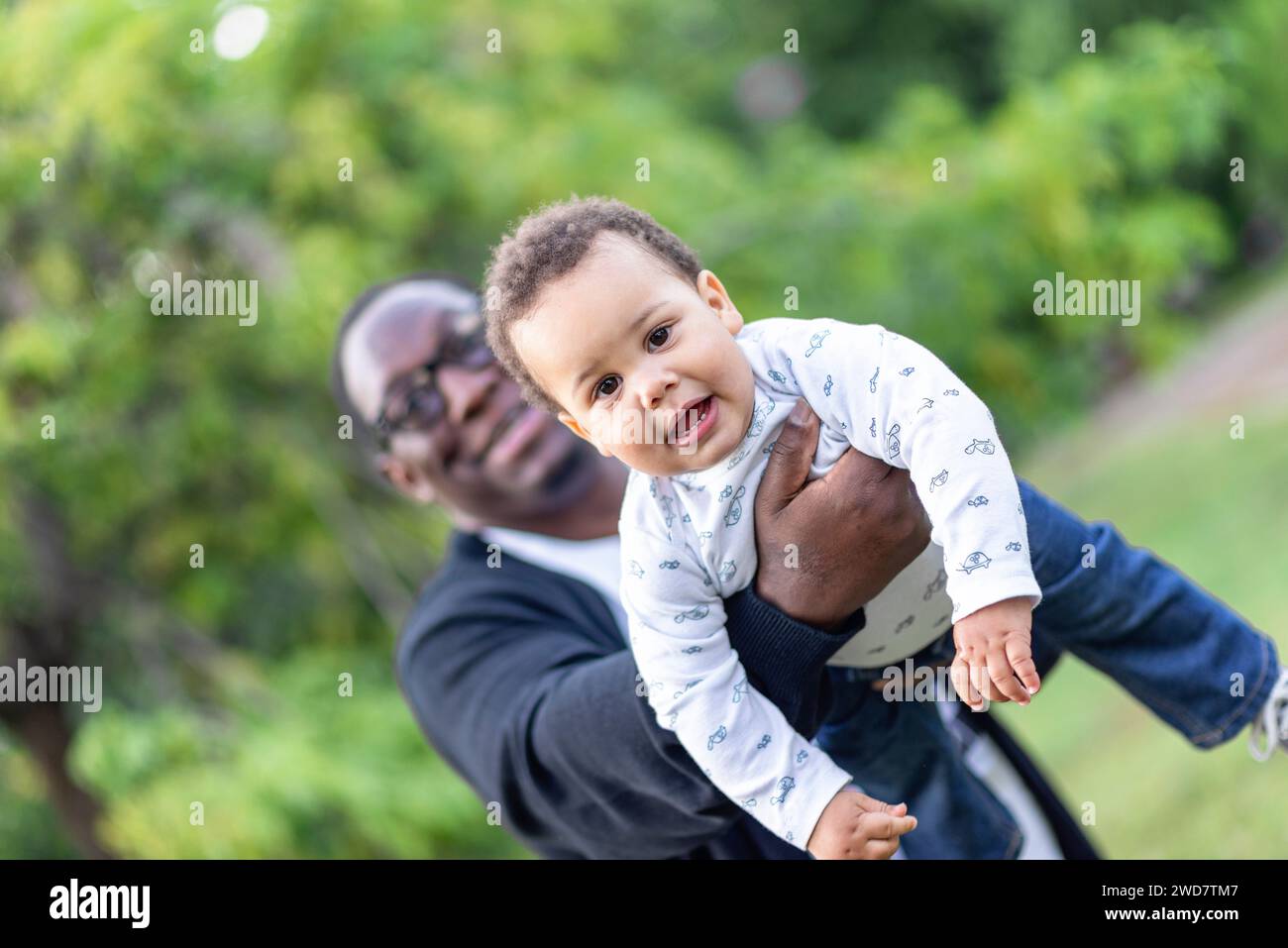 Vater kuschelt niedlichen birassischen Baby-Jungen, kleiner afroamerikanischer Junge, der die Kamera ansieht, wie er sich draußen mit Dad verbündet Stockfoto