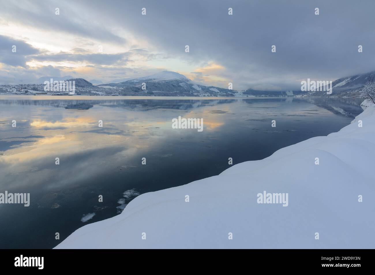 Das Bild fängt eine ruhige Winterszene mit einem glatten, spiegelähnlichen Fjord ein, der die sanften Farben des Abendhimmels in schneebedeckter Umgebung reflektiert. Stockfoto