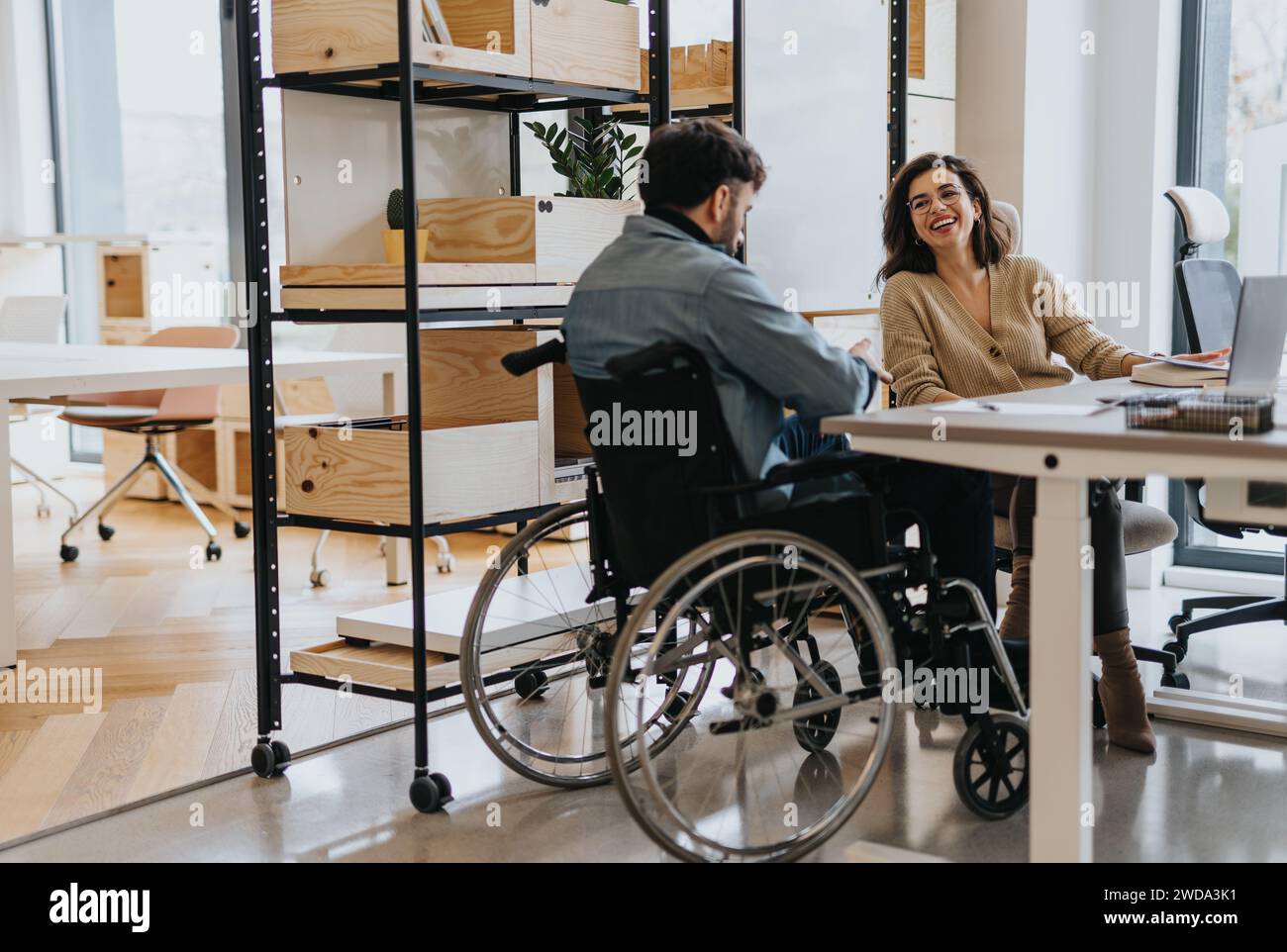 Wunderschöne Frau mittleren Alters, die mit ihrem behinderten männlichen Kollegen im Rollstuhl in einem modernen Kreativbüro über die Arbeit spricht. Stockfoto