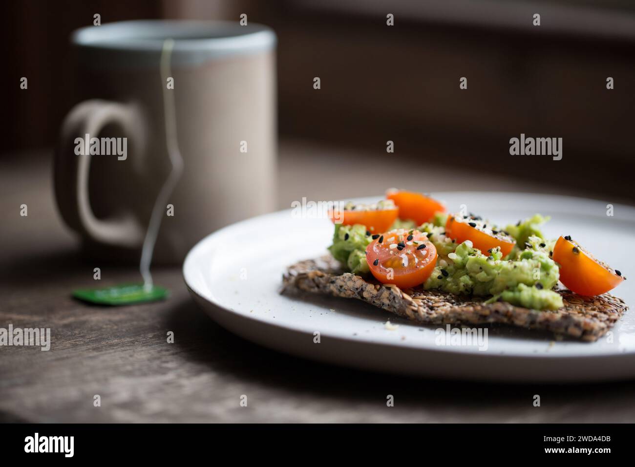 Leinsamen Cracker mit zerstoßener Avocado, Kirschtomaten und Samen, Tasse Tee im Hintergrund, einfache gesunde Frühstücksidee. Stockfoto