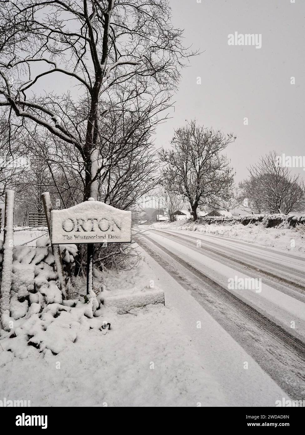 Schnee fällt in einer winterlichen Szene in Nordengland mit einem Ortsschild mit angrenzender Straße und Bäumen unter einer Schneedecke. Stockfoto