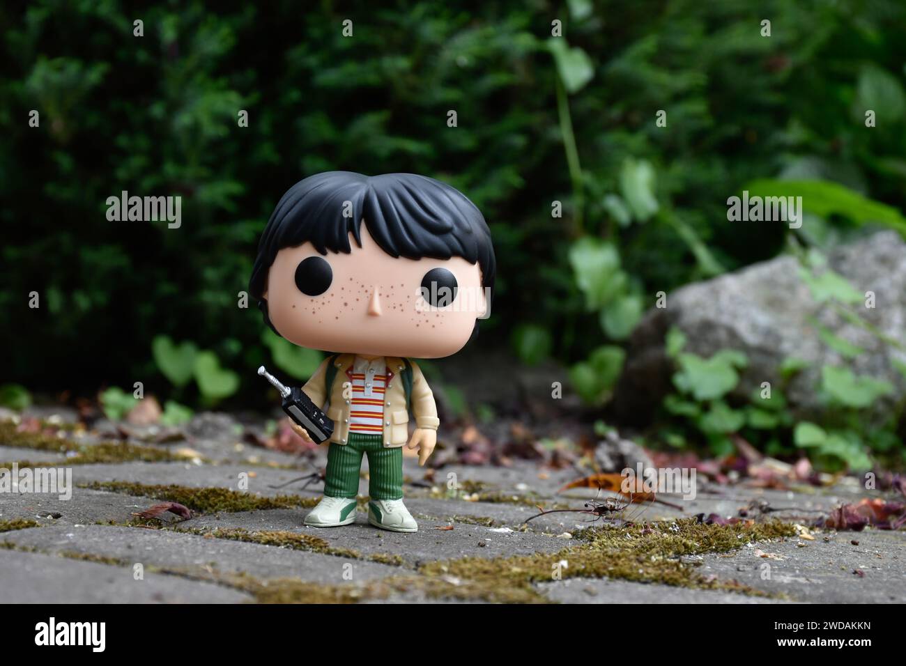 Funko Pop Actionfigur von Mike mit Walkie-Talkie aus der beliebten Horror-TV-Serie Stranger Things. Grüne Blätter, moosige Steinstraße, Frühlingsgarten. Stockfoto