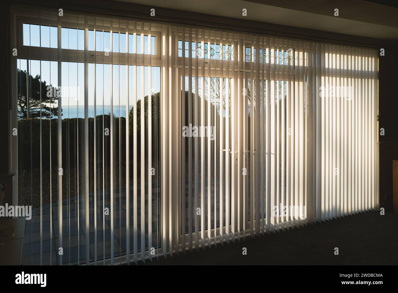 Sonnenlicht, das durch weiße vertikale Jalousien in voller Länge vor drei Glasschiebetüren strahlt, die zu einer Terrasse, Garten und Meer führen Stockfoto