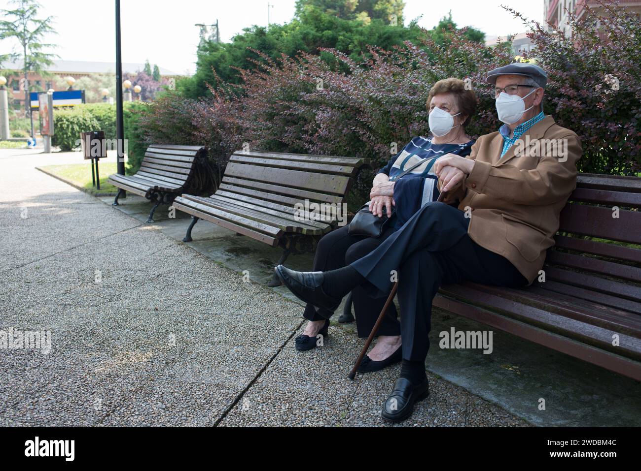 Seniorenpaar mit Masken auf einer Stadtbank. Mann, der einen Gehstock benutzt Stockfoto