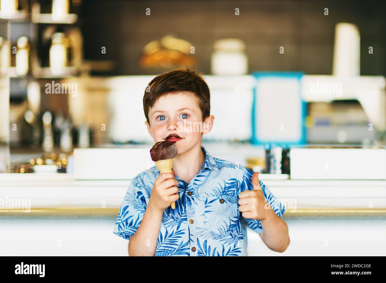 Lustiger Junge isst Schokoladeneis in der Eisdiele, großer Daumen hoch Stockfoto