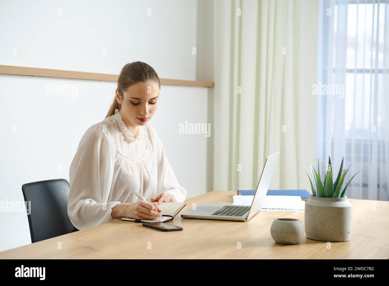 Konzentrierte junge Frau in weißer Bluse, die Notizen macht, während sie im Büro mit Laptop arbeitet Stockfoto