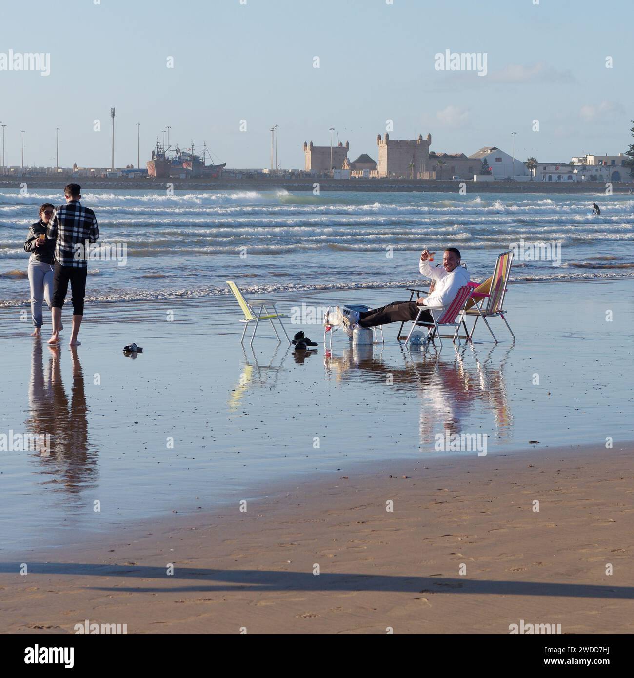 Freunde am Strand, während einer von ihnen auf einem Liegestuhl im flachen Wasser sitzt, mit dem Fort in der Medina dahinter in Essaouira, Marokko, 19. Januar 2024 Stockfoto