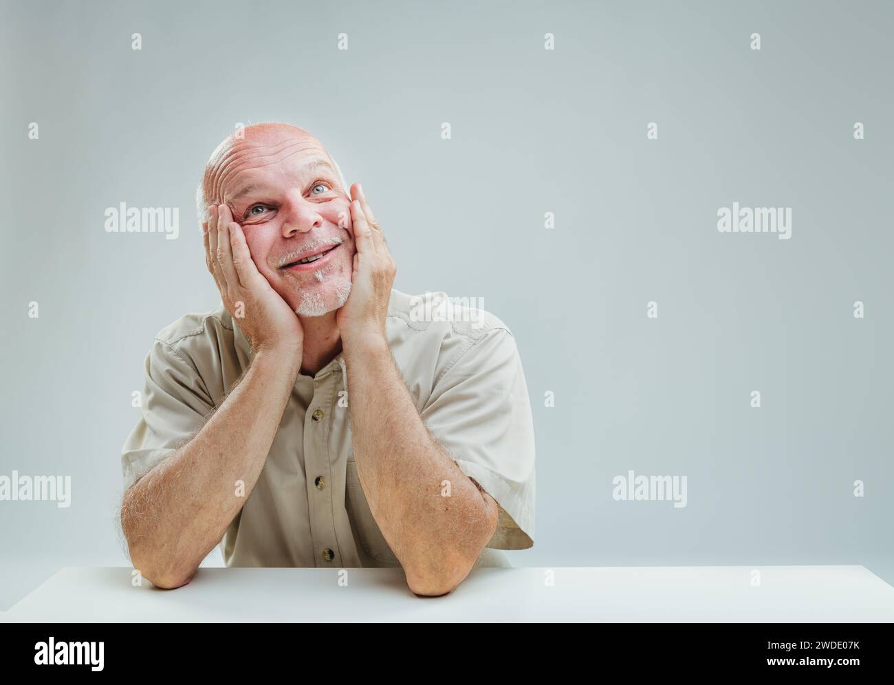 Ein älterer Mann träumt mit einem zarten Lächeln, Hände schröpfen die Wangen in einer Geste der Zufriedenheit Stockfoto