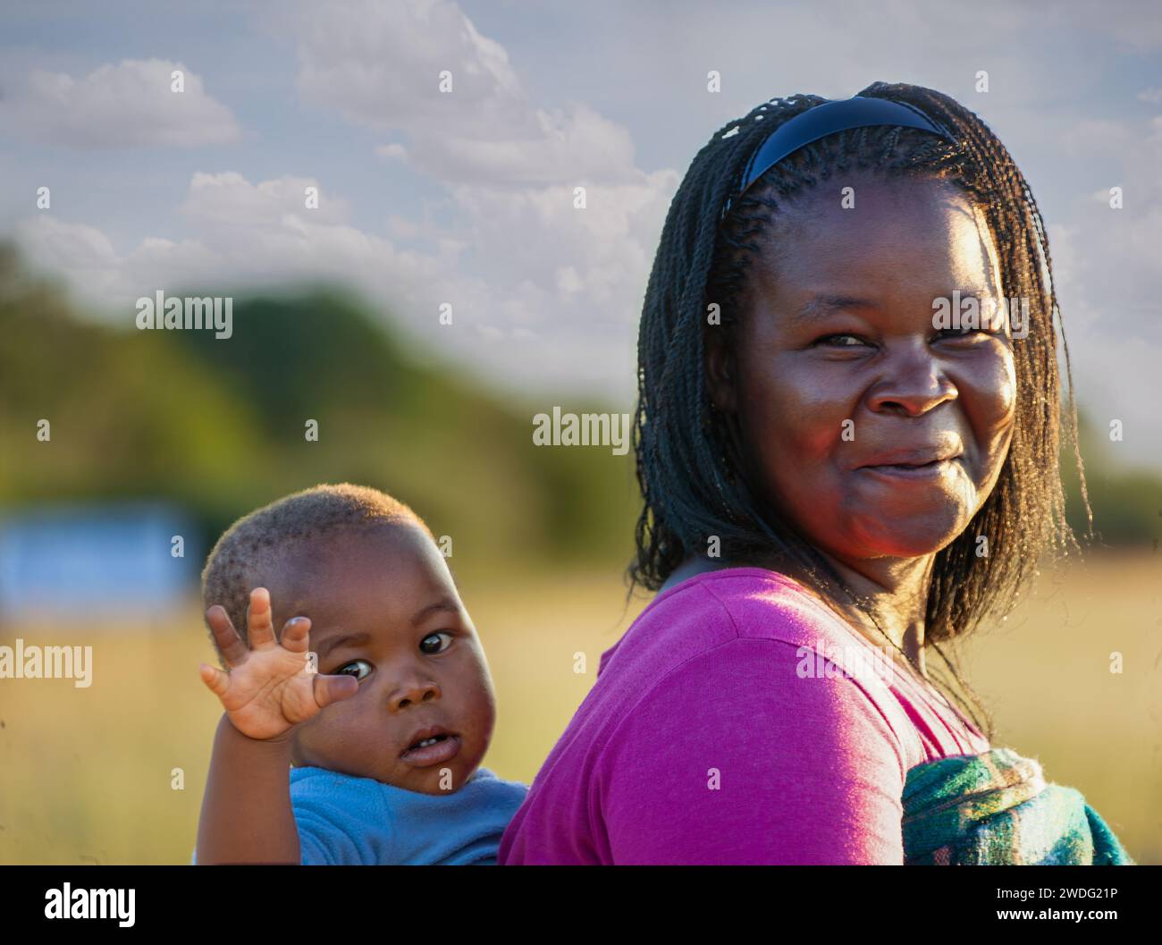 Eine afrikanische Dorffrau mit Zöpfen trägt das Kind auf dem Rücken auf traditionelle Weise in einer Decke Stockfoto