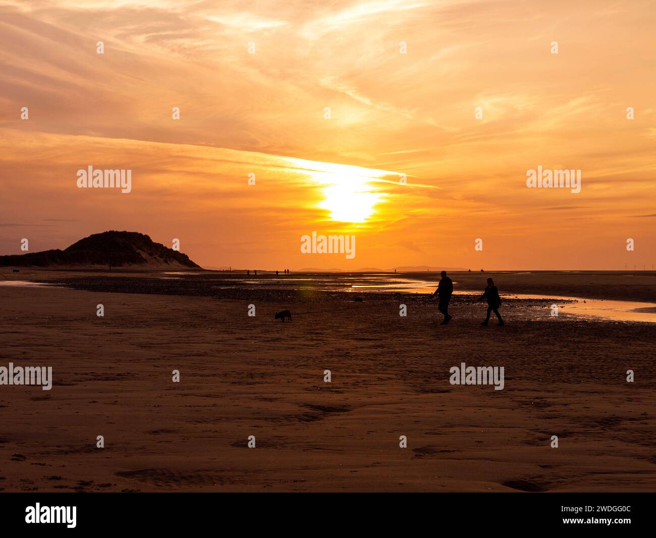 Silhouette-Paar, das einen Hund bei Sonnenuntergang am Talacre Beach bei Ebbe spaziert, Talacre, Flintshire, Wales Stockfoto