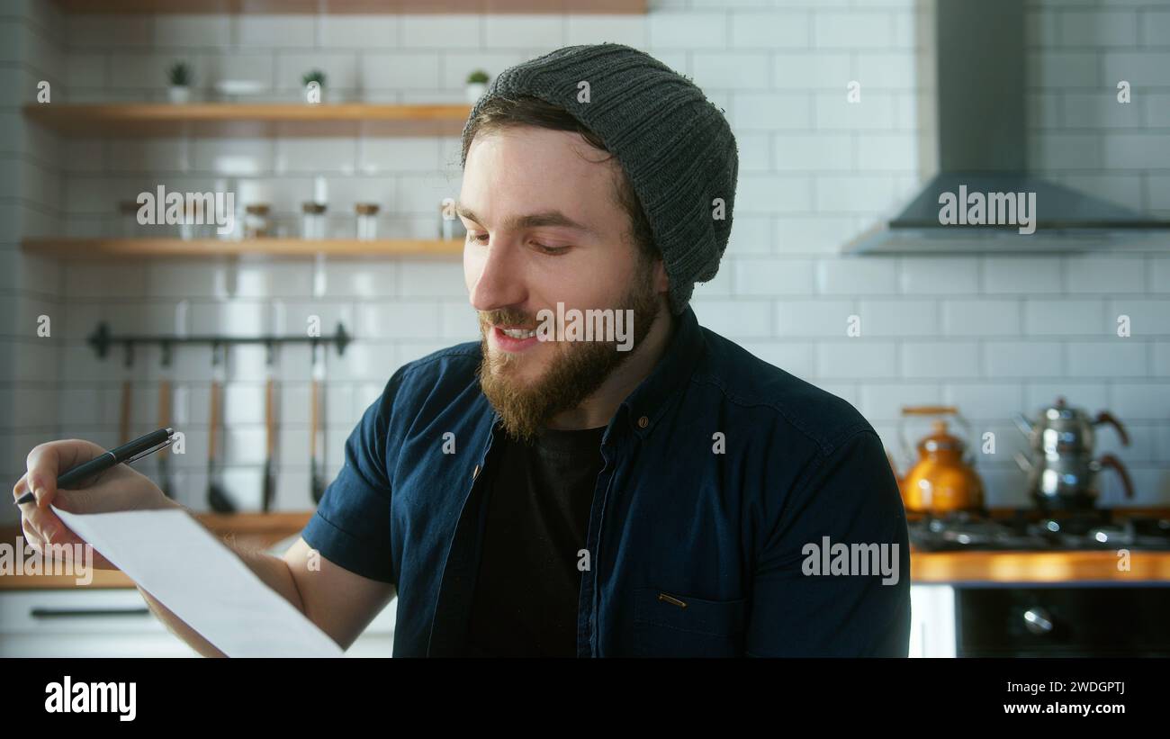 Junger erwachsener Mann mit Beanie, der zu Hause in der modernen Küche sitzt, spricht und Berichte in Videokonferenzgesprächen zeigt. Selbstbewusste Männer haben Videokonferenz Stockfoto